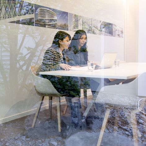 wo women sitting next to each other behind a laptop in a white meeting room.